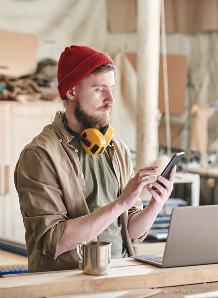 Ein junger Handwerker mit einem Smartphone in der Hand an einer Werkbank sitzend mit einem Laptop drauf